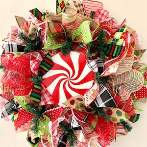 May include: A Christmas wreath decorated with red, green, white, and brown ribbon, mesh, and a peppermint swirl design. The wreath is decorated with gingerbread men and other festive details.