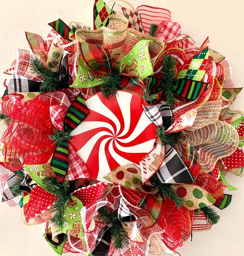 May include: A Christmas wreath decorated with red, green, white, and brown ribbon, mesh, and a peppermint swirl design. The wreath is decorated with gingerbread men and other festive details.