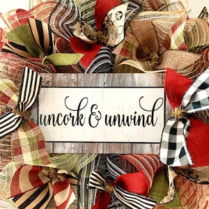 May include: A burlap wreath with red, black, and white checkered ribbon, green and brown plaid ribbon, and a sign that says "uncork & unwind".