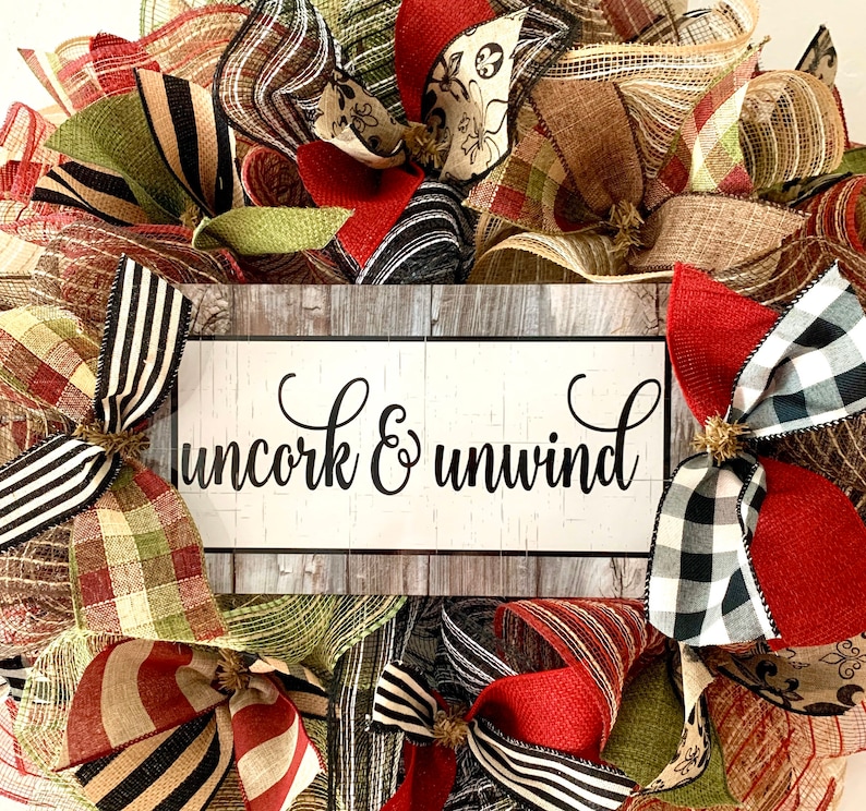 May include: A burlap wreath with red, black, and white checkered ribbon, green and brown plaid ribbon, and a sign that says "uncork & unwind".