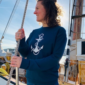 Könnte beinhalten: Eine Frau trägt ein marineblaues Sweatshirt mit einem weißen Anker-Motiv auf der Vorderseite. Sie steht auf einem Holzdeck eines Bootes und hält ein Seil.