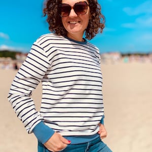 Könnte beinhalten: Eine Person trägt ein weiß-blau gestreiftes Sweatshirt mit blauen Bündchen und einem blauen Kragen, kombiniert mit einer blauen Jeans. Die Person trägt auch eine Sonnenbrille. Der Hintergrund ist ein Strand mit einem strahlend blauen Himmel.