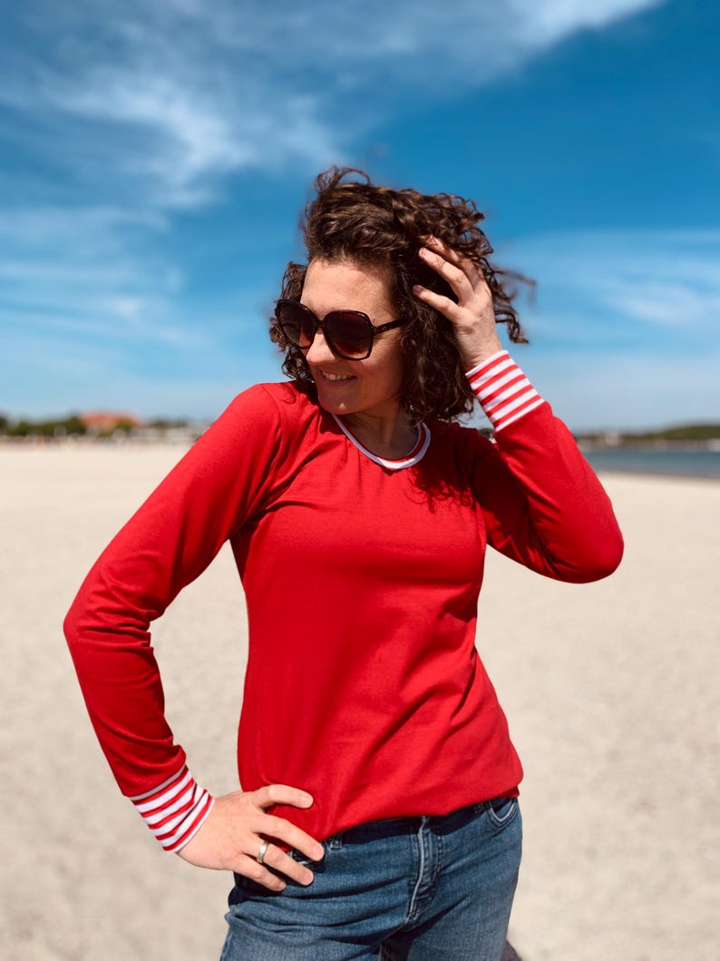 May include: A red long-sleeve shirt with white and red striped cuffs and a white collar. The person is wearing blue jeans and sunglasses. The background is a beach with a blue sky.