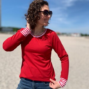 May include: A red long-sleeve shirt with white and red striped cuffs and neckline. The shirt is paired with blue jeans and sunglasses. The background is a beach with a blue sky.