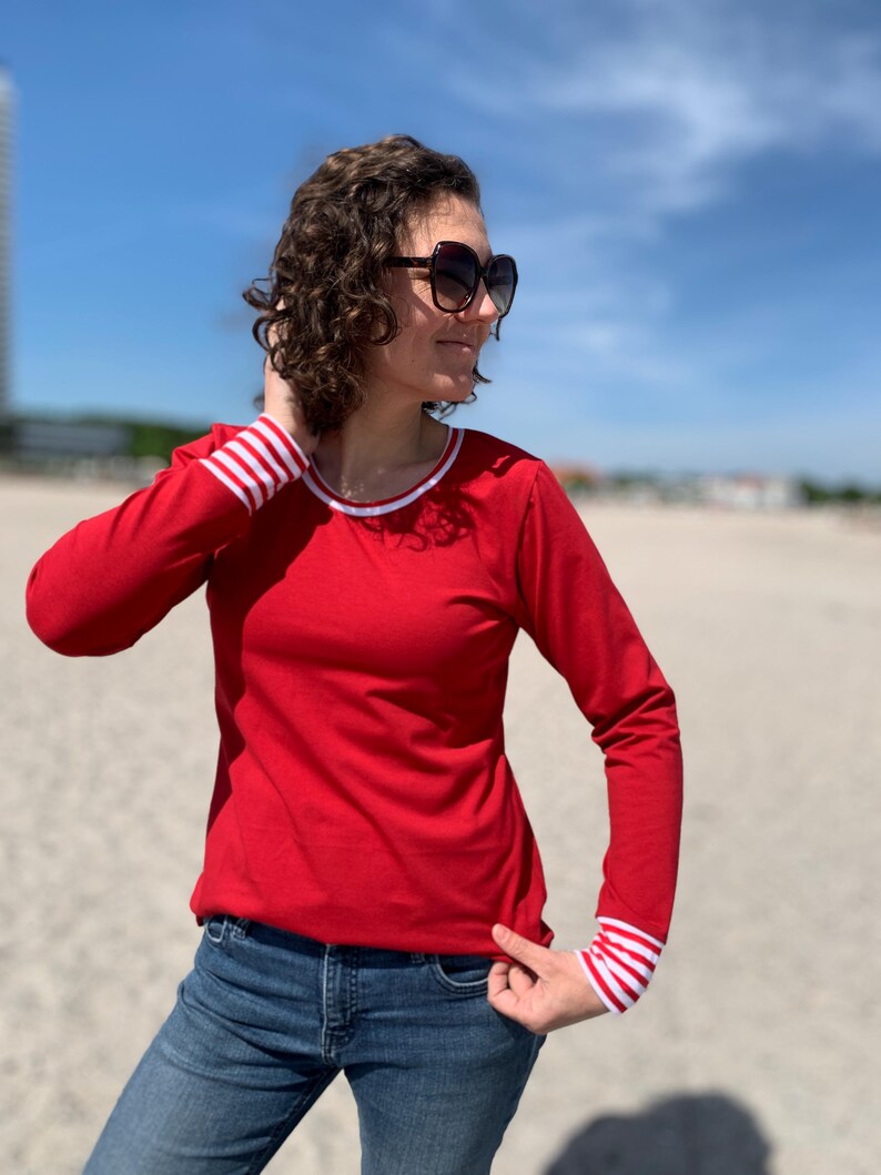 May include: A red long-sleeve shirt with white and red striped cuffs and neckline. The shirt is paired with blue jeans and sunglasses. The background is a beach with a blue sky.