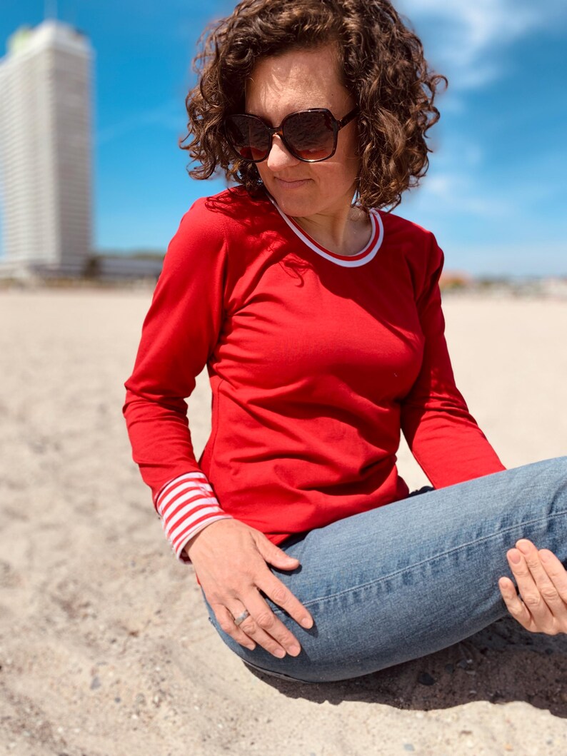 May include: A red long-sleeve shirt with white and red striped cuffs and a white collar trim. The person is wearing blue jeans and sunglasses. The background includes a beach and a tall building.
