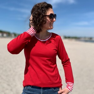 May include: A red long-sleeve shirt with white and red striped cuffs and neckline trim. The shirt is paired with blue jeans. The person is wearing sunglasses.