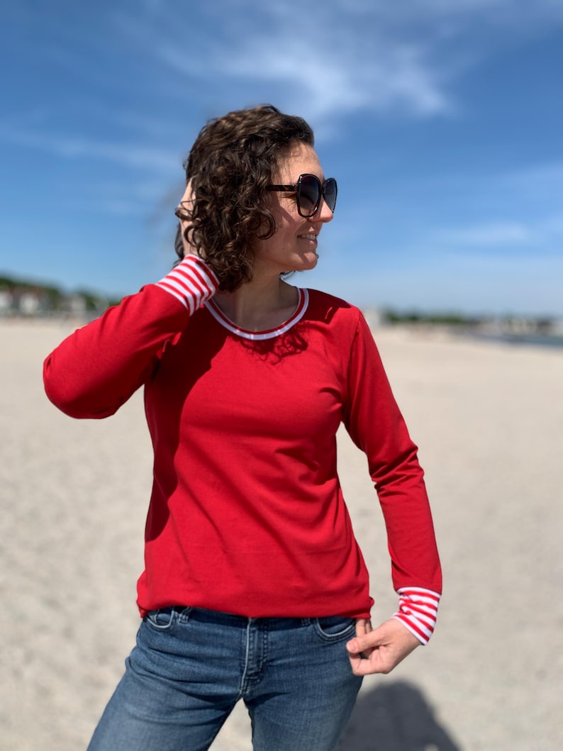 May include: A red long-sleeve shirt with white and red striped cuffs and neckline trim. The shirt is paired with blue jeans. The person is wearing sunglasses.