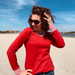 May include: A red long-sleeve shirt with white and red striped cuffs and a white collar. The person is wearing blue jeans and sunglasses. The background is a beach with a blue sky.