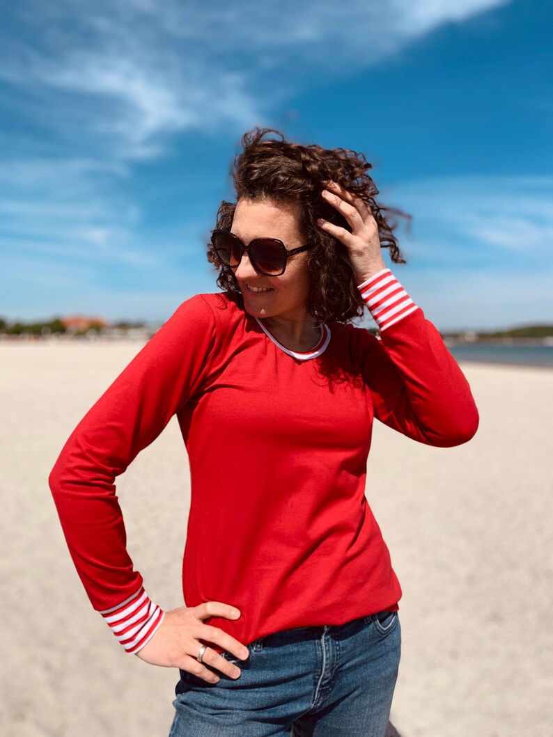 May include: A red long-sleeve shirt with white and red striped cuffs and a white collar. The person is wearing blue jeans and sunglasses. The background is a beach with a blue sky.