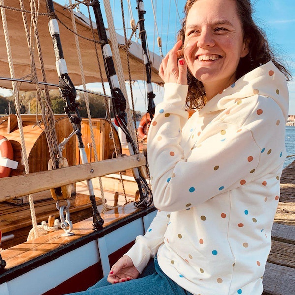 May include: A woman wearing a white hoodie with colorful polka dots, blue jeans, and a big smile. She is sitting on a wooden boat deck.