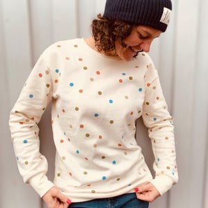 Könnte beinhalten: Ein cremefarbener Pullover mit einem Tupfenmuster in Blau, Braun und Orange. Der Pullover hat lange Ärmel und einen Rundhalsausschnitt.