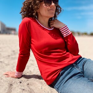 May include: A person wearing a red long-sleeve shirt with white and red striped cuffs and a white collar. The person is wearing blue jeans and sunglasses. The background is a beach with a blue sky.