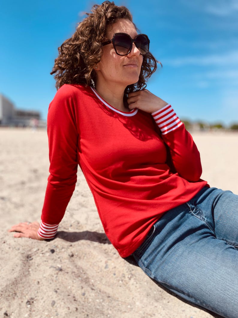 May include: A person wearing a red long-sleeve shirt with white and red striped cuffs and a white collar. The person is wearing blue jeans and sunglasses. The background is a beach with a blue sky.