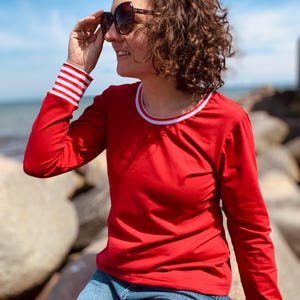 May include: A red long-sleeve shirt with white and red striped cuffs and a white collar trim. The person is wearing blue jeans and sunglasses. The background shows a blue sky and ocean.