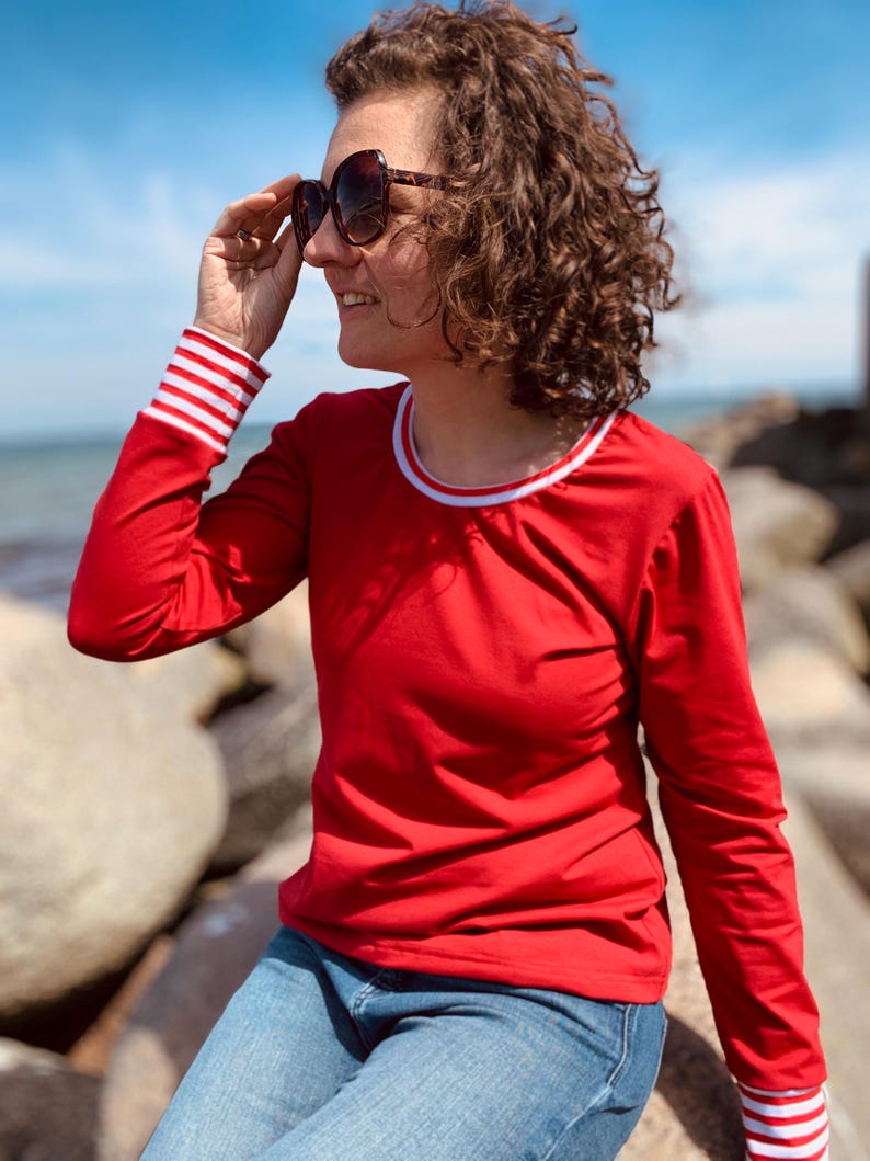 May include: A red long-sleeve shirt with white and red striped cuffs and a white collar trim. The person is wearing blue jeans and sunglasses. The background shows a blue sky and ocean.