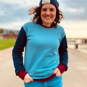 Könnte beinhalten: Eine Frau trägt eine blaue Mütze mit dem Wort "Moin", einen hellblauen und marineblauen Farbblock-Sweatshirt und eine blaue Jeans. Das Sweatshirt hat bordeauxfarbene Bündchen und einen Halsausschnitt. Die Frau lächelt.