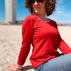 May include: A red long-sleeve shirt with white trim on the collar and cuffs. The cuffs have a white and red striped pattern. The person is wearing blue jeans and sunglasses. The background includes a beach and a tall white building.