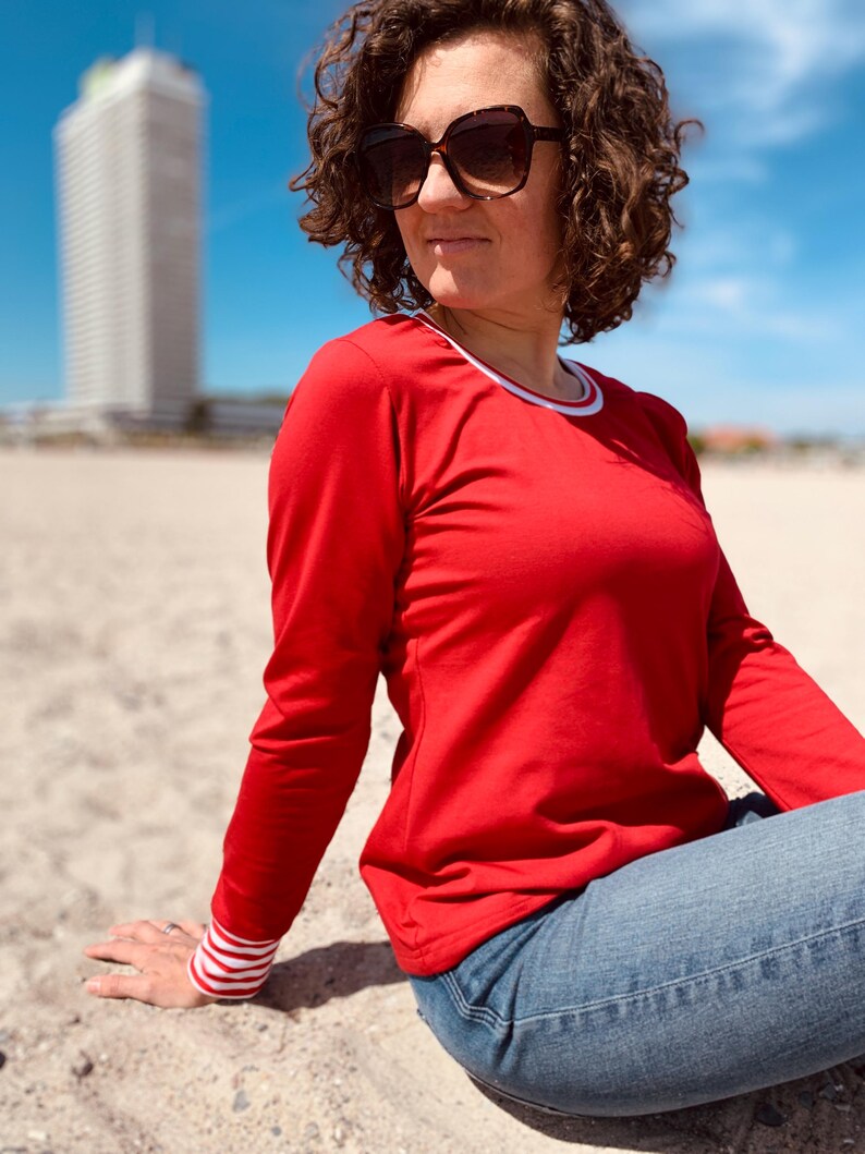 May include: A red long-sleeve shirt with white trim on the collar and cuffs. The cuffs have a white and red striped pattern. The person is wearing blue jeans and sunglasses. The background includes a beach and a tall white building.