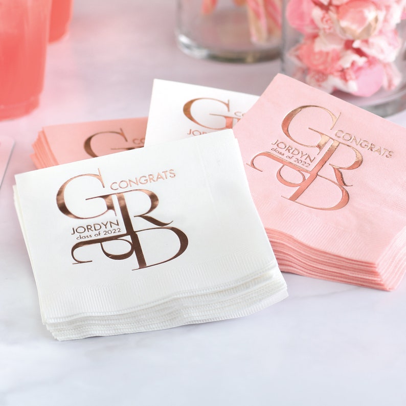 personalized graduation beverage napkins featuring a large GRAD monogram design in shiny foil. White and blush pink napkins are shown stacked on a party table with rose gold foil text reading “Congrats” and the graduate’s name, with space to personalize the graduation year. These custom napkins are perfect for graduation parties and celebrations and are available in nine napkin colors with multiple foil color options for the printed design.