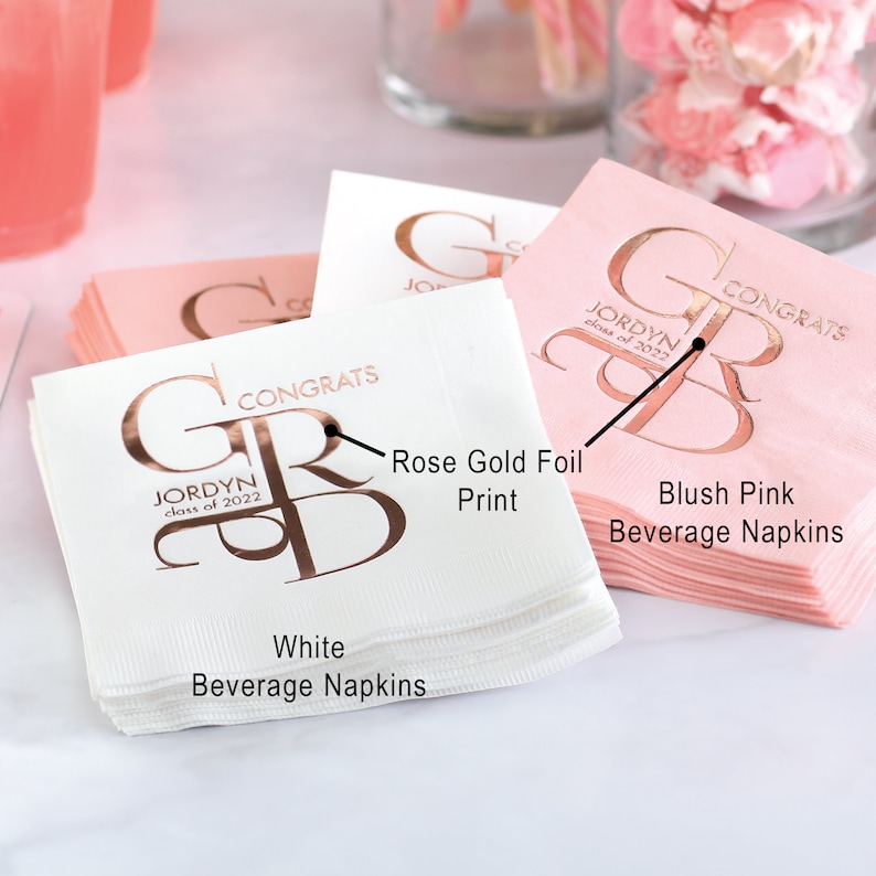 personalized graduation beverage napkins featuring a large GRAD monogram design in shiny foil. White and blush pink napkins are shown stacked on a party table with rose gold foil text reading “Congrats” and the graduate’s name, with space to personalize the graduation year. These custom napkins are perfect for graduation parties and celebrations and are available in nine napkin colors with multiple foil color options for the printed design.