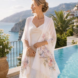 Puede incluir: Un chal rectangular blanco y ligero con un delicado estampado floral multicolor en tonos rosa, morado y verde en los bordes, combinado con un vestido lencero largo de satén color crema sin mangas con escote tipo capucha.