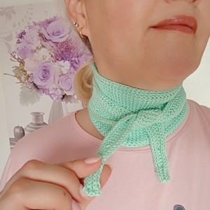 Puede incluir: Un calentador de cuello de punto verde claro con un lazo atado en la parte delantera.