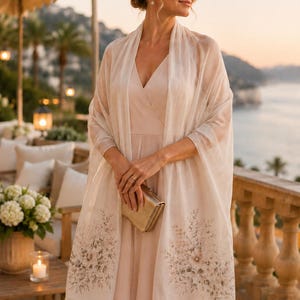Puede incluir: Un chal de color beige claro con un delicado estampado floral en tonos suaves en ambos extremos, acabado con un sutil detalle de flecos. La tela rectangular se coloca sobre los hombros, mostrando una textura suave y semitransparente.