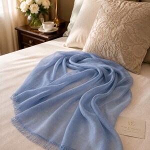 Puede incluir: Una bufanda rectangular de color azul claro con un delicado fleco en los bordes. Junto a la bufanda hay una peque&ntilde;a tarjeta rectangular con el texto "PE PURE ELEGANCE TIMELESS BEAUTY" impreso en fuente oscura sobre fondo claro.
