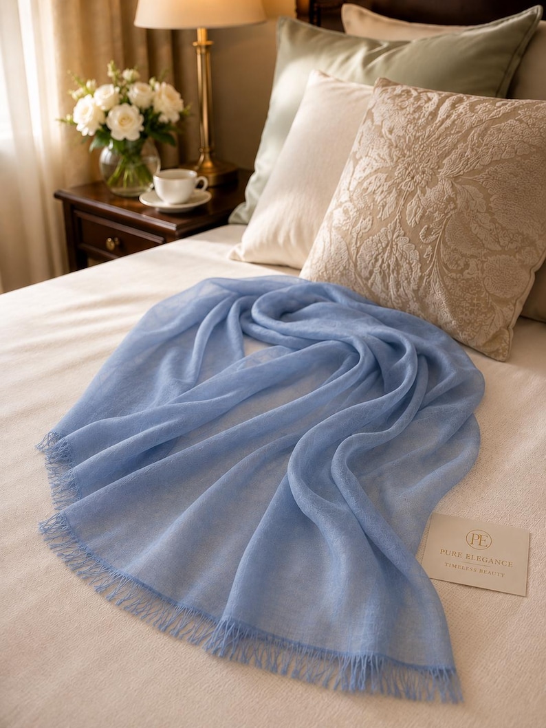 Puede incluir: Una bufanda rectangular de color azul claro con un delicado fleco en los bordes. Junto a la bufanda hay una peque&ntilde;a tarjeta rectangular con el texto "PE PURE ELEGANCE TIMELESS BEAUTY" impreso en fuente oscura sobre fondo claro.