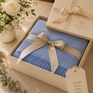 Puede incluir: Una manta azul claro doblada con bordes de flecos, presentada en una caja de regalo color crema con una cinta de sat&eacute;n. La tapa tiene el texto PURE ELEGANCE TIMELESS BEAUTY. Una etiqueta dice A Beautiful Gift for a Special Occasion.