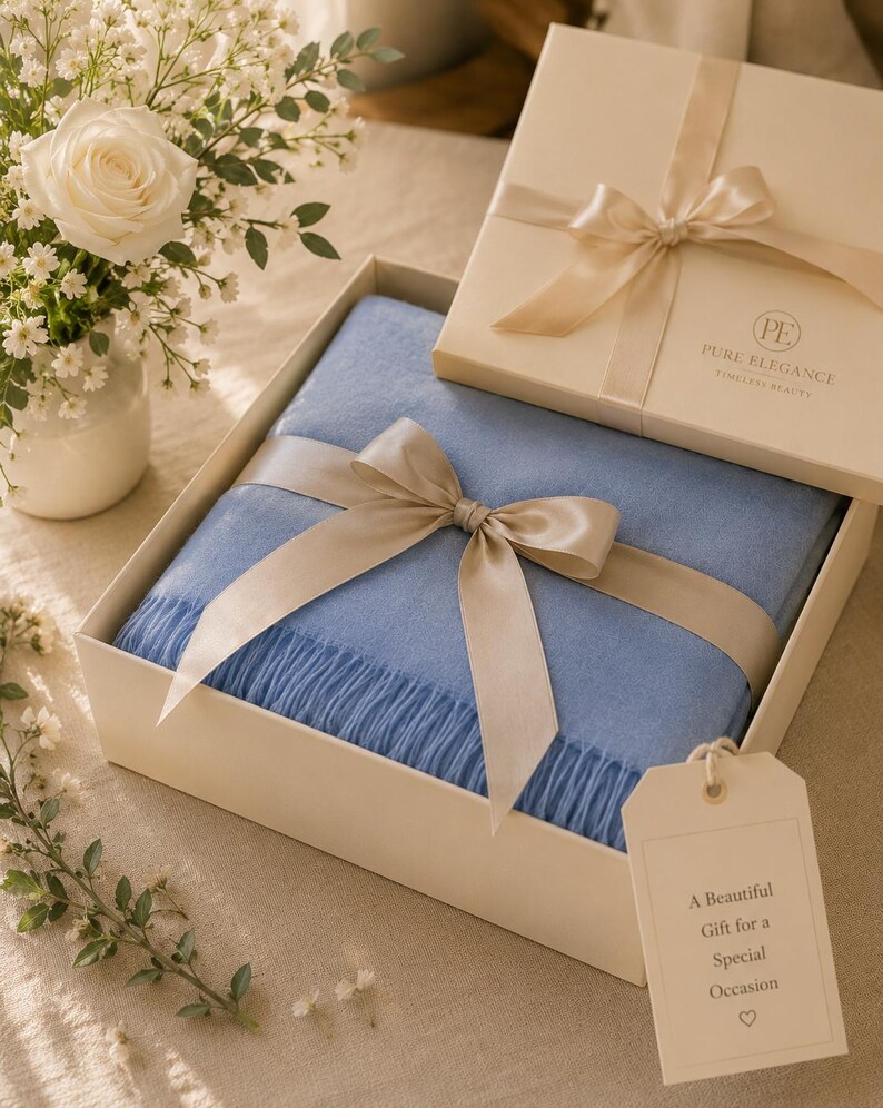 Puede incluir: Una manta azul claro doblada con bordes de flecos, presentada en una caja de regalo color crema con una cinta de sat&eacute;n. La tapa tiene el texto PURE ELEGANCE TIMELESS BEAUTY. Una etiqueta dice A Beautiful Gift for a Special Occasion.