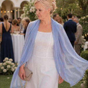 Puede incluir: Un vestido blanco sin mangas con escote cuadrado combinado con un chal rectangular azul claro transparente con un delicado borde de flecos. Un peque&ntilde;o bolso de mano rectangular de color beige con una fina cadena dorada se sostiene en la mano.