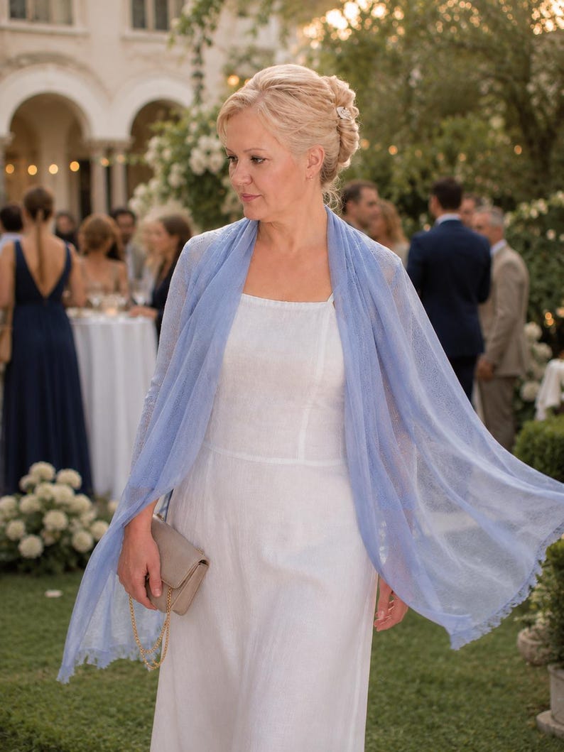 Puede incluir: Un vestido blanco sin mangas con escote cuadrado combinado con un chal rectangular azul claro transparente con un delicado borde de flecos. Un peque&ntilde;o bolso de mano rectangular de color beige con una fina cadena dorada se sostiene en la mano.