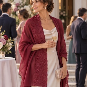 Puede incluir: Un chal transparente de color burdeos intenso con un intrincado borde bordado de flores, drapeado sobre un vestido color crema con cuello tipo cascada. La tela ligera tiene una textura suave y sirve como accesorio elegante para eventos formales.