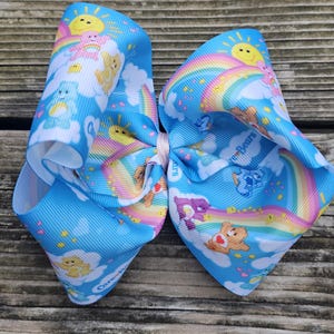 May include: A large, light blue hair bow featuring a repeating pattern of Care Bears, rainbows, and clouds. The bow is tied in the centre with a light pink ribbon. The Care Bears are in various poses.