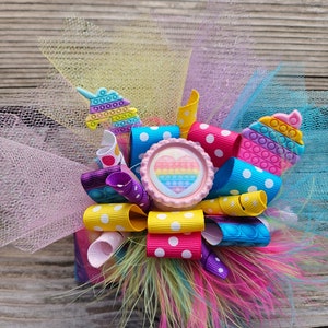 May include: A colourful hair bow featuring a rainbow heart, polka dot ribbon and a unicorn charm. The bow is made with tulle and feathers.