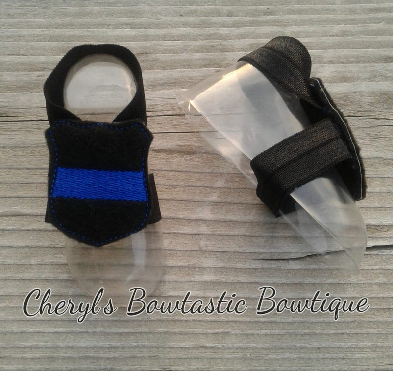 May include: A pair of black dog booties with a blue embroidered line on one bootie. The booties are on a wooden surface.