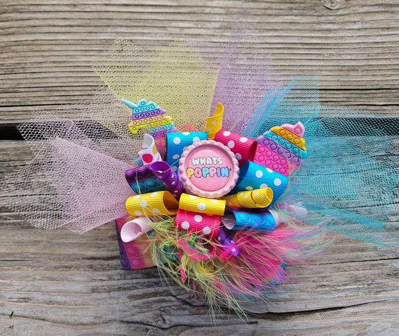 May include: A colourful hair bow featuring a pink button with the text "Whats Poppin'" and a blue and yellow pop it toy. The bow is made with tulle and ribbon.