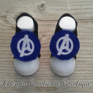 May include: A pair of baby sandals with a blue and white Avengers logo on each strap. The sandals are black and white with a white sole.