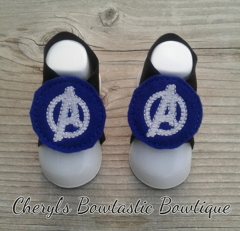 May include: A pair of baby sandals with a blue and white Avengers logo on each strap. The sandals are black and white with a white sole.