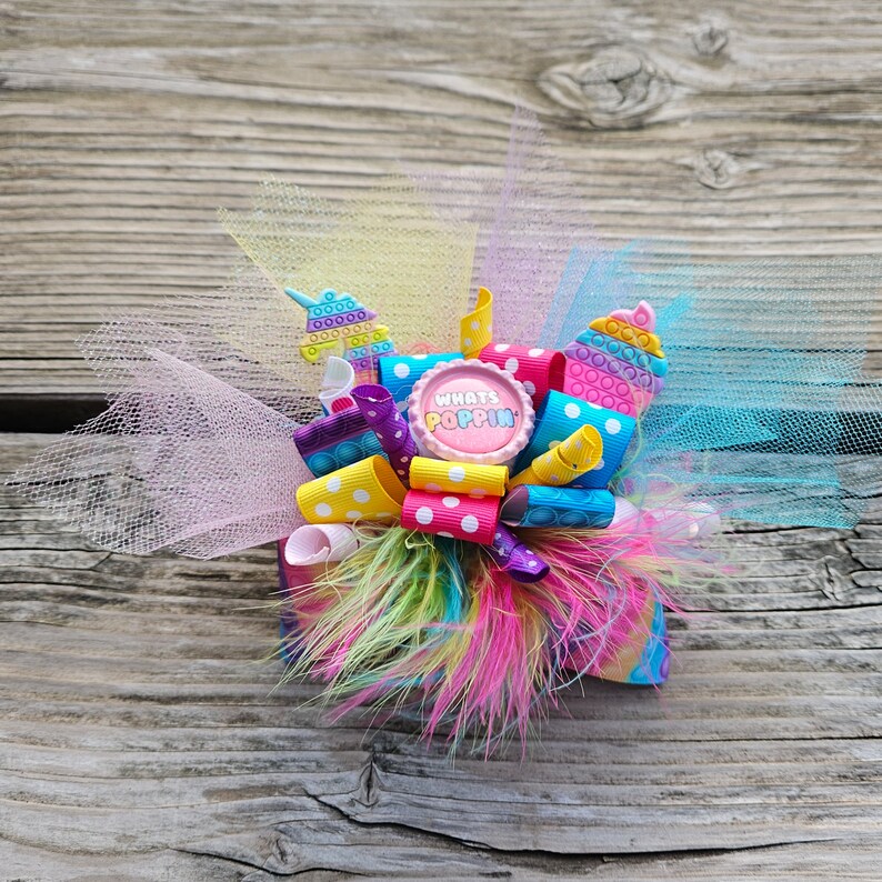 May include: A colourful hair bow featuring a pink button with the text "Whats Poppin". The bow is made from tulle, ribbon and feathers. It is decorated with colourful pop-it toys.