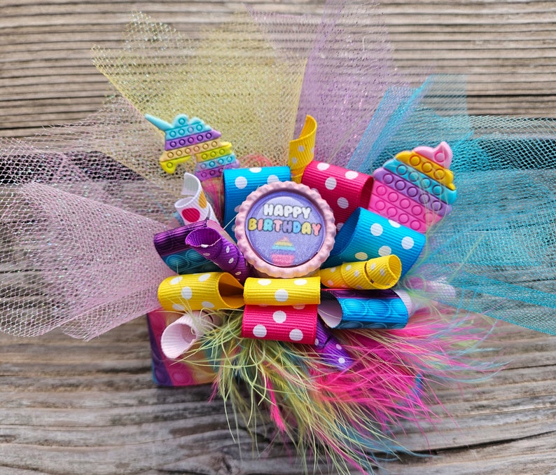 May include: A colourful birthday hair bow featuring a purple cupcake charm with the words "Happy Birthday". The bow is made with yellow, pink, blue and purple ribbon with white polka dots. The bow is also decorated with a unicorn charm made of colourful plastic.