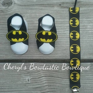 May include: A pair of black baby sandals with yellow Batman logos. The sandals are on a wooden surface. A black suspender with yellow Batman logos is also pictured.
