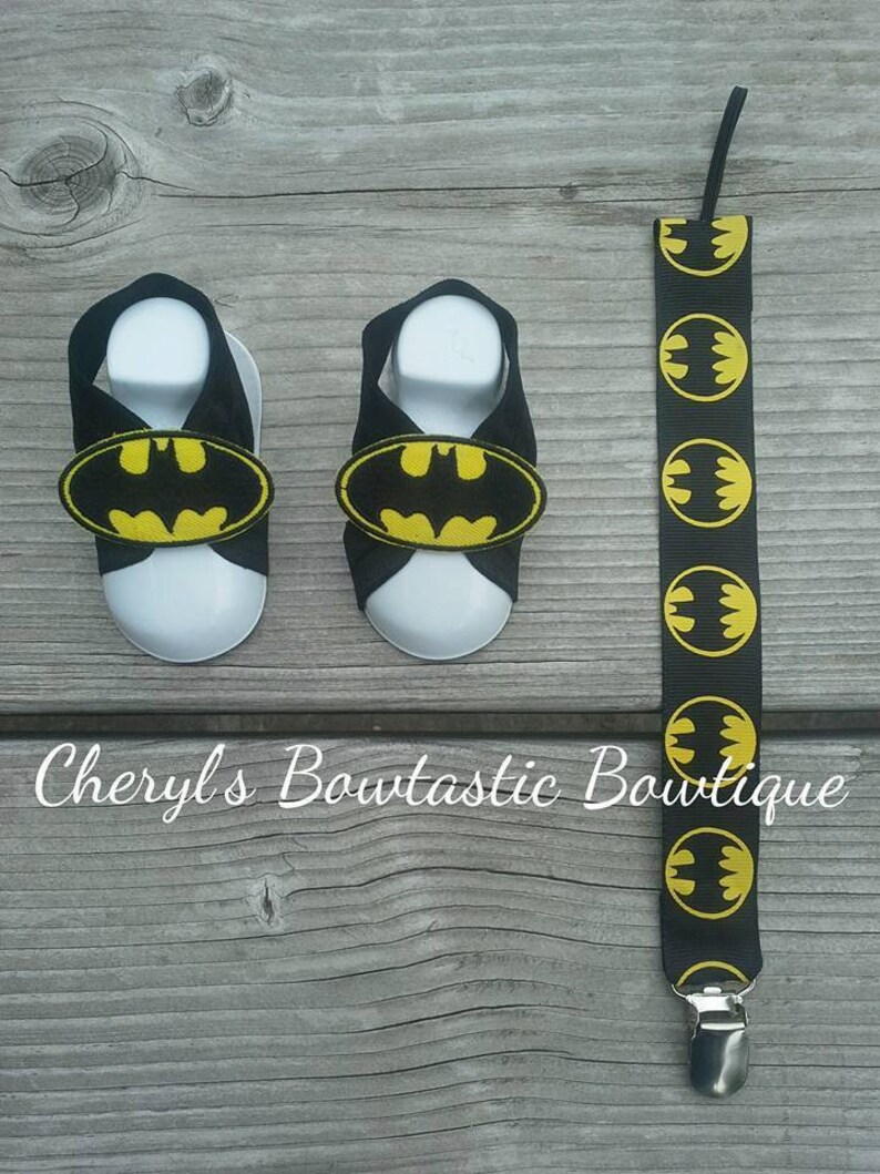 May include: A pair of black baby sandals with yellow Batman logos. The sandals are on a wooden surface. A black suspender with yellow Batman logos is also pictured.