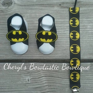 May include: A pair of black baby sandals with yellow Batman logos and a black and yellow Batman logo patterned suspender clip.