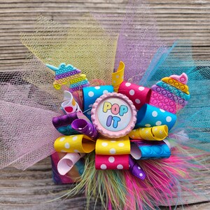 May include: A colourful hair bow with a pink button that says "Pop It" in the centre. The bow is made of yellow, pink, blue, and purple ribbon with white polka dots. The bow is surrounded by pink, yellow, and blue tulle.