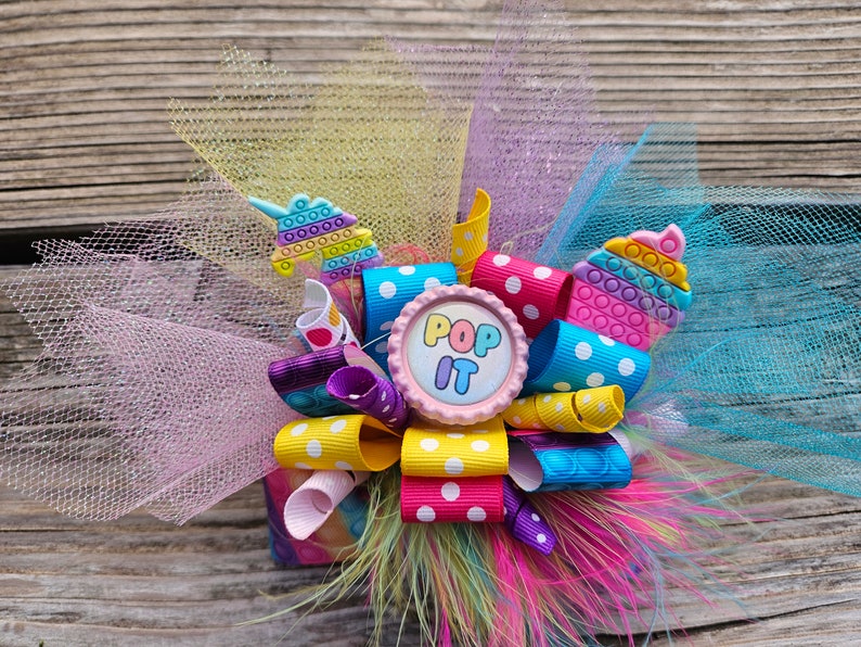 May include: A colourful hair bow with a pink button that says "Pop It" in the centre. The bow is made of yellow, pink, blue, and purple ribbon with white polka dots. The bow is surrounded by pink, yellow, and blue tulle.