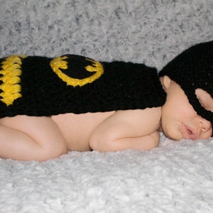 May include: A baby wearing a black and yellow crocheted Batman costume, sleeping on a white fuzzy blanket.