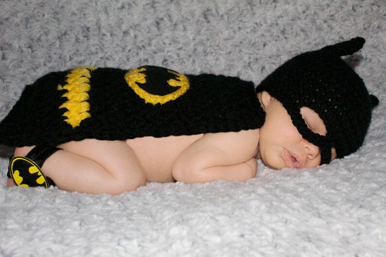 May include: A baby wearing a black and yellow crocheted Batman costume, sleeping on a white fuzzy blanket.